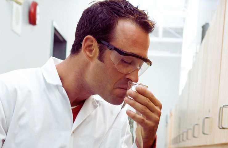 uomo con occhiali protettivi annusa una provetta e fa una faccia disgustata
