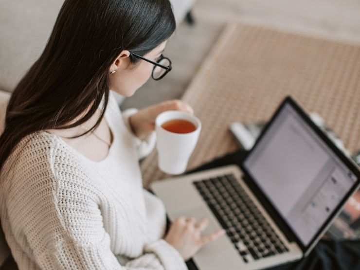 Una ragazza è seduta sul tappeto e dal computer invia cv mentre beve una tazza di caffè