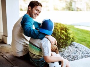 Un padre e un figlio seduti sui gradini di casa dopo aver giocato a pallone. Stanno parlando e si sorridono.
