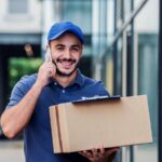 Un corriere consegna un pacco sorridente mentre parla al telefono col cliente.